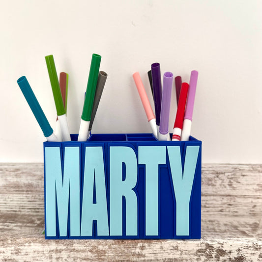 Blue pencil holder with the name 'MARTY' holding colorful markers on a wooden surface.