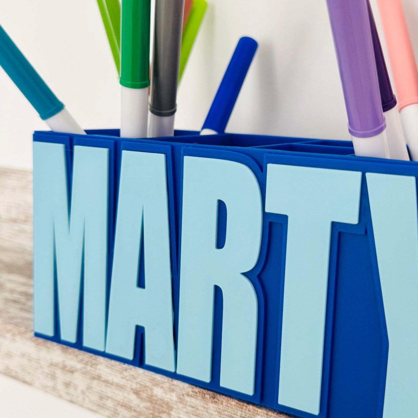 Blue pen holder with the name 'MARTY' holding colorful markers on a wooden surface.